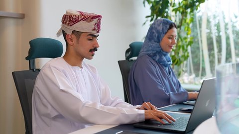 الحوارات والنقاشات ضمن الفريق, مفهوم الشراكة والتعاون, بيئة العمل العماني ، رجل عربي خليجي عماني يرتدي الدشداشة و العمامة يجلس فى مكتبة برفقة زميلته يقوم بالعمل على الحاسب المحمول ، ايماءات الاندماج و التركيز