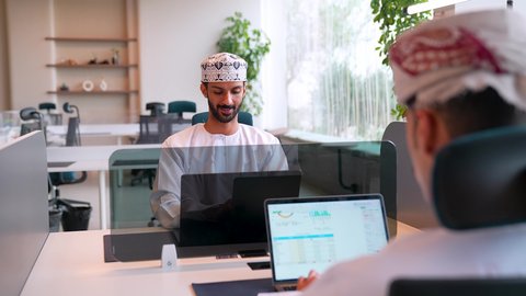 بيئة العمل المريحة في الشركات العمانية، إنجاز المهام المكتبية باستخدام الحاسب المحمول , رجل عربي خليجي عماني مبتسم يرتدي الدشداشة و الكمة يقوم بالعمل على الابتوب يجلس امام زميله, دمج التقنية والتكنولوجيا مع الحياة العملية