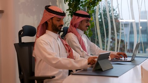 أداء المهام والاعمال المكتبية داخل مقر الشركة، رجل عربي خليجي سعودي مبتسم يرتدي الثوب التقليدي والشماغ يجلس أمام مكتبه يقوم بالعمل على الحاسب المحمول برفقة زميله، دمج التقنية والتكنولوجيا في الحياة العملية، العمل في مكاتب وشركات المملكة العربية السعودية