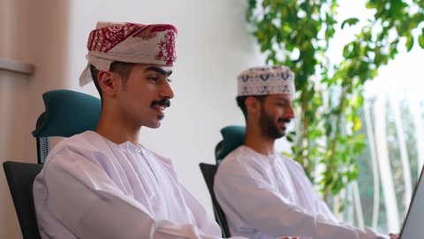 إدارة شؤون الأعمال وتنظيمها، تصويرلرجل عربي خليجي عماني يرتدي الزي التقليدي يقوم بالعمل على الحاسوب داخل المكتب برفقة زميله، العمل لتحقيق الاهداف والاستراتيجيات، مهن ووظائف ادارية مكتبية, استخدام الأجهزة التكنولوجية الحديثة والمتطورة