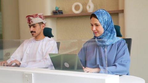 دمج العمل مع التقنية والتكنولوجيا الحديثة والمتطورة، ادارة وانجاز المهام باستخدام جهاز تقني حديث ومتطور, تصويرلامراة عربيه خليجيه عمانية محجبه ترتدي العباءة تعمل على الحاسوب المحمول في مقر الشركة برفقة زميلها، مهن ووظائف مكتبية اماراتية