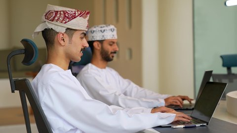 إدارة شؤون الأعمال وتنظيمها عبر الاجهزة التقنية، مهن ووظائف ادارية مكتبية، دمج العمل مع التقنية والتكنولوجيا، رجلان عربيان حليجيان عمانيان يرتدي الزي التقليدي يقومان بالعمل على الحاسوب المحمول داخل المكتب