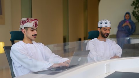 النقر على لوحة المفاتيح، مهن ووظائف مكتبيه عمانية ، إدارة سير الأعمال عبر الحاسوب المحمول، مفهوم الإشراف على العمل، زميلان عربيان خليجيان عمانيان يرتديان الزي التقليدي يجلسان على المكتب يقومان بالعمل على الابتوب داخل الشركة