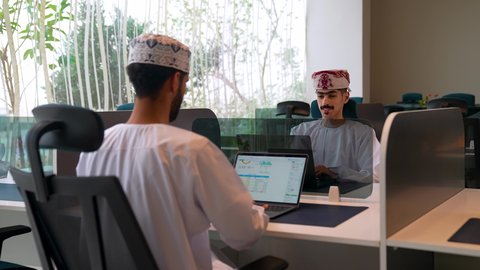 مهن ووظائف مكتبيه عمانية، الاستفادة من التقنيات الحديثة في القطاع المكتبي والإداري، إدارة سير الأعمال عبر الحاسوب المحمول، مفهوم الإشراف على العمل، زميلان عربيان خليجيان عمانيان يرتديان الزي التقليدي يجلسان على المكتب يقومان بالعمل على الابتوب داخل الشركة