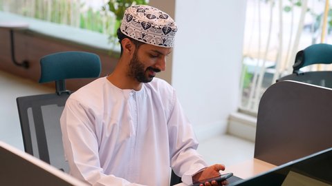 تصفح مواقع التواصل الاجتماعي، الدردشة مع الأهل والأصدقاء، استخدام جهاز تقني حديث, دمج التكنولوجيا المتطورة بالحياة اليومية، تصويرمقرب لرجل عربي خليجي عماني يرتدي الدشداشة والكمة يجلس على مكتبة و يستخدم الهاتف المحمول