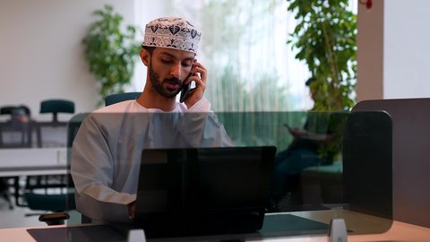 التواصل مع الأهل والأصدقاء عبر الهاتف المحمول ، الرد على الاتصالات والمكالمات، تصوير لرجل عربي خليجي عماني يرتدي الدشداشة و الكمه يجلس على مكتب فى مقر الشركة و يقوم بالعمل على الحاسب المحمول ، استخدام الأجهزة الحديثة والمتطورة