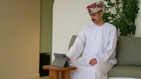 The use of modern and advanced technology for learning, the use of a tablet to accomplish work, an Arab Omani man wearing traditional attire browsing the internet on a tablet, using a modern technological device.