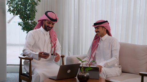 Using the mobile phone to take commemorative selfies, discussing plans and explaining work tasks, developing businesses and companies, dialogues and discussions within the team, Arab Saudi Gulf colleagues in a meeting, wearing traditional Saudi attire, gestures of happiness and joy.