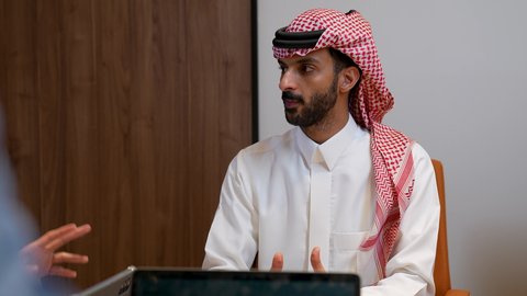 النقاشات والحوارات ضمن الفريق، اكتساب الخبرة وتوسيع الخطط المهنية، تصوير مقرب لرجل عربي خليجي سعودي يرتدي الزي التقليدي و الشماغ يقوم  بتدريب الموظفين على سياسات ومهام العمل لتحقيق الأهداف في غرفة الاجتماع داخل مقر الشركة