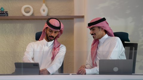 التفاهم والتعاون بين الفريق، تدريب الموظفين على سياسات ومهام العمل لتحقيق الأهداف في غرفة الاجتماع داخل مقر الشركة، بيئة العمل المريحة في الشركات السعودية، استخدام الأجهزة والتقنيات الحديثة، رجل أعمال عربي خليجي سعودي يرتدي الزي التقليدي و الشماغ  يقوم بمناقشة المشروع مع زميله