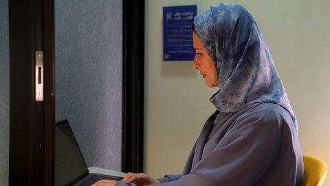 استخدام الأجهزة والتقنيات الحديثة، مهن و أعمال إدارية مكتبية، النقر على  لوحه المفاتيح، تصوير من الجانب لامراة عربيه خليجيه سعودية ترتدي العباءة تجلس على مكتب فى مقر الشركة و تقوم بالعمل على الحاسب المحمول