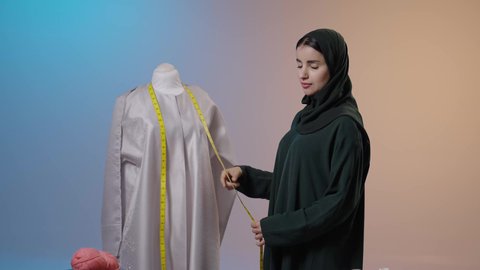 النظر الى الكاميرا بايماءات السعادة و السرور ، أدوات التطريز والخياطة، إعداد موديلات نسائية مميزة، أخذ القياسات المناسبة باستخدام الشريط المتري , بورتريه لمصممة أزياء عربية خليجية سعودية ترتدي العباءة السوداء والحجاب تقف أمام المانيكان