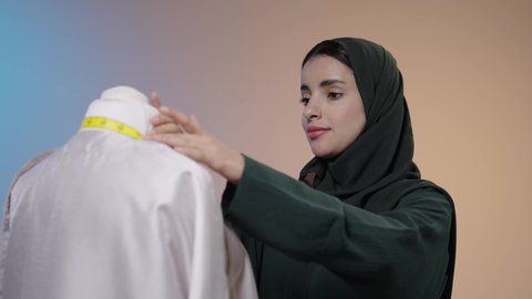 أخذ القياسات المناسبة باستخدام الشريط المتري , خياطة و تفصيل الملابس النسائية , بورتريه لمصممة أزياء عربية خليجية سعودية ترتدي العباءة السوداء والحجاب تقف أمام المانيكان، أدوات و معدات الخياطة