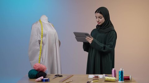 التسويق عبر الإنترنت من خلال التابلت، تصفح مواقع السوشيال ميديا، استخدام جهاز تقني حديث، استعمال أدوات ومعدات الخياطة، إتقان صنع الملابس السعودية، بورتريه لخياطة عربية خليجية سعودية ترتدي العباءة السوداء تقف امام المانيكان تستخدم التابلت