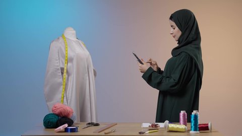 Using modern technical devices, mobile electronic marketing, browsing social media sites, using sewing tools and equipment, mastering the making of Saudi clothing, a portrait of a Gulf Arab Saudi seamstress wearing a black abaya standing in front of a mannequin using a mobile phone.