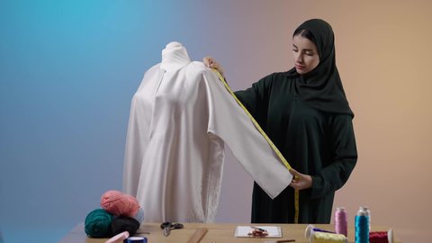 أدوات التطريز والخياطة، إعداد موديلات نسائية مميزة، أخذ القياسات المناسبة باستخدام الشريط المتري , بورتريه لمصممة أزياء عربية خليجية سعودية ترتدي العباءة السوداء والحجاب تقف أمام المانيكان
