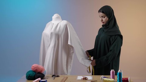 معدات وخامات الخياطة، إعداد موديلات نسائية مميزة، أخذ القياسات المناسبة باستخدام الشريط المتري , بورتريه لمصممة أزياء عربية خليجية سعودية ترتدي العباءة السوداء والحجاب تقف أمام المانيكان