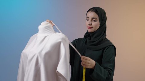 أخذ القياسات المناسبة باستخدام الشريط المتري , خياطة و تفصيل الملابس النسائية , بورتريه لمصممة أزياء عربية خليجية سعودية ترتدي العباءة السوداء والحجاب تقف أمام المانيكان، أدوات و معدات الخياطة