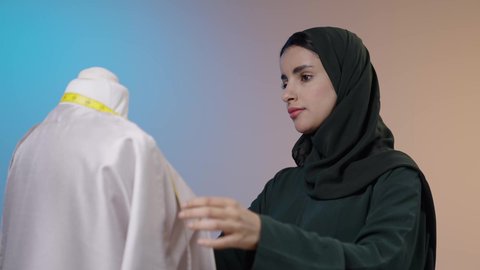 تفصيل فساتين وأزياء نسائية، مهن و أعمال الخياطة و التصميم, بورتريه مقرب لمصممه أزياء عربية خليجية سعودية ترتدي الحجاب و العباءة السوداء تقف أمام المانيكان تأخذ القياسات بالشريط المتري , خلفية ملونه