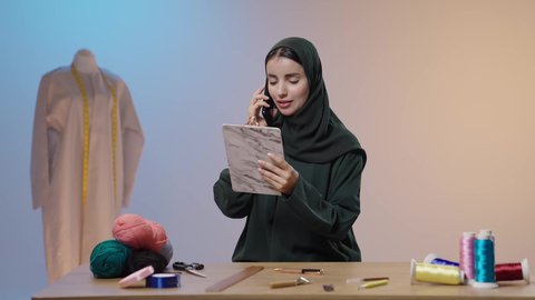 Using sewing supplies and tools, mastering the making of Saudi clothing, communicating via mobile phone, exchanging news and conversations, using advanced and modern technologies, a portrait of a Saudi Gulf Arab seamstress wearing a black abaya, using a mobile phone and holding a document holder in her hands, with a colorful background.