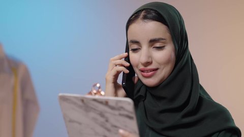 Making a phone call, exchanging news and conversations, using advanced and modern technologies, using sewing tools and equipment, mastering the making of Saudi clothing, a close-up portrait of a Saudi Gulf Arab seamstress wearing a black abaya, using a mobile phone and holding a document holder in her hands, with a colorful background.