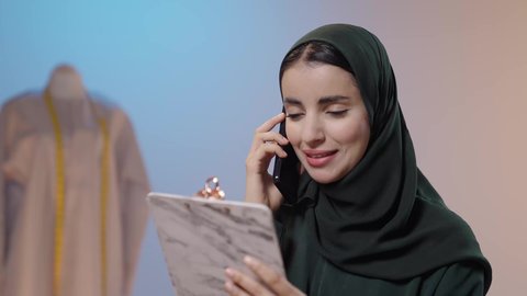 تبادل الأخبار والأحاديث، استخدام التقنيات المتطورة والحديثة، إجراء مكالمة هاتفية، استعمال أدوات ومعدات الخياطة، إتقان صنع الملابس السعودية، بورتريه مقرب لخياطة عربية خليجية سعودية ترتدي العباءة السوداء تستخدم الهاتف المحمول و تمسك بيديها حافظه الاوراق، خلفية ملونة