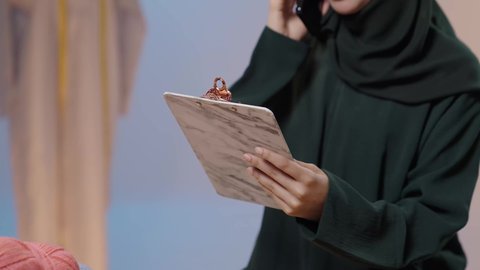 استخدام التقنيات المتطورة والحديثة، تبادل الأخبار والأحاديث، إجراء مكالمة هاتفية، استعمال أدوات ومعدات الخياطة، إتقان صنع الملابس السعودية، بورتريه مقرب لخياطة عربية خليجية سعودية ترتدي العباءة السوداء تستخدم الهاتف المحمول و تمسك بيديها حافظه الاوراق، خلفية ملونة