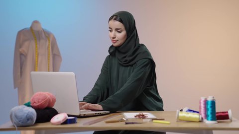 مهن و حرف يدوية، استخدام الأجهزة التقنية المتطورة , خياطة و تفصيل الملابس النسائية , بورتريه لمصممة أزياء عربية خليجية سعودية ترتدي العباءة السوداء و الحجاب تحمل جهاز الحاسوب بيديها ,خلفية ملونه