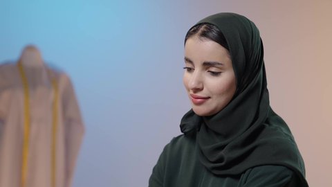 مهن و أعمال الخياطة و التصميم, بورتريه مقرب لمصممه أزياء عربية خليجية سعودية ترتدي الحجاب و العباءة السوداء تتأمل في ابتكار موديلات جديدة، خلفية ملونه