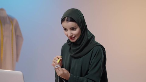 الإبداع في تقديم أزياء جديدة ، ممارسة مهنة الخياطة، تصوير مقرب لامراة عربية خليجية سعودية ترتدي الحجاب و العباءة السوداء تستخدم إبرة الكروشيه وخيوط الصوف تجلس امام الحاسب المحمول ، أعمال تقليدية شعبية, فنون الخياطة و التصميم ، ايماءات السعادة و السرور