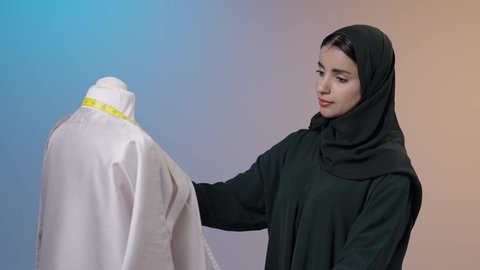 مهن و أعمال الخياطة و التصميم , تفصيل الملابس النسائية , بورتريه لمصممة أزياء عربية خليجية سعودية ترتدي العباءة السوداء والحجاب تقف أمام المانيكان تأخذ القياسات بالشريط المتري , خلفية ملونه