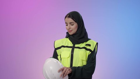 المشاريع الهندسية ومخططات الإعمار, بورتريه مقرب لفتاة عربية خليجية سعودية مبتسمة ترتدي سترة الحماية والخوذة تقف بأيدي متقاطعة وتنظر إلى الكاميرا بايماءات السعادة و السرور, العمل في القطاع الهندسي, تحقيق حلم النجاح والوصول لمهنة الهندسة بتعابير الفخر، خلفية ملونه