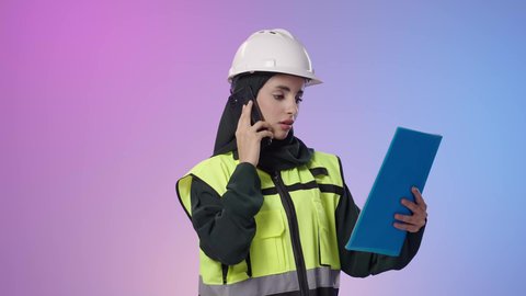 استخدام الأجهزة التقنية الحديثة، مفهوم الهندسة والانشاءات المعمارية، بورتريه مقرب لمهندسة عربية خليجية سعودية ترتدي سترة الخاصة وخوذة الحماية تمسك بيدها الهاتف المحمول و الاوراق تقوم بمتابعة الأعمال، النظر الى شيء ما ، خلفية ملونه