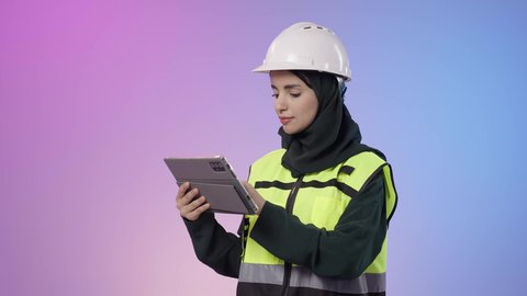 التطور والتحول التقني الحديث، مهن ووظائف نسائية سعودية، دمج البيئة العملية و الهندسه مع التقنية و التكنولوجيا, بورتريه  مقرب لامراة عربيه خليجيه سعودية محجبه ترتدي الخوذة وسترة الحماية تحمل بيديها جهاز التابلت و تقوم بالنظر الى شيء ما، خلفية ملونه