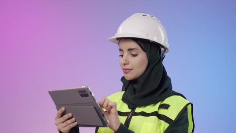 متابعة سير العمل بالمشاريع الهندسية، دمج البيئة العملية و الهندسه مع التقنية و التكنولوجيا, بورتريه  مقرب لامراة عربيه خليجيه سعودية محجبه ترتدي الخوذة وسترة الحماية تحمل بيديها جهاز التابلت و تقوم بالنظر الى شيء ما، خلفية ملونه