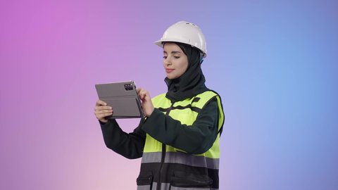 Standing confidently with arms crossed, merging the work environment and engineering with technology and tech, electronic engineering plans and designs, a portrait of a Saudi Arabian Gulf woman wearing a hijab, helmet, and safety vest, holding a tablet in her hands, with a colorful background.