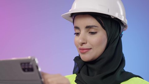 دمج البيئة العملية و الهندسه مع التقنية و التكنولوجيا, المخططات و التصماميم الهندسية الالكترونية, بورتريه مقرب لامراة عربيه خليجيه سعودية محجبه ترتدي الخوذة وسترة الحماية تحمل بيديها جهاز التابلت، مفهوم الهندسة و الإنشاءات المعمارية، خلفية ملونه