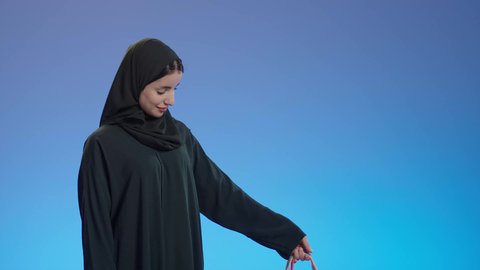 عروض وتخفيضات موسميه، الاستمتاع بالتبضع فى السعودية، التسوق في المراكز التجاريه، بورتريه لإمرأة عربيه خليجية سعوديه محجبة ترتدي عباءه سوداء تحمل بيدها أكياس التسوق, إيماءات تدل على السعاده، خلفية ملونه