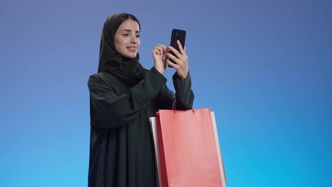 حقائب تسوق ملونه عروض وتخفيضات موسميه، بورتريه لإمرأة عربيه خليجية سعوديه ترتدي العباءة السوداء والحجاب تحمل بيدها أكياس التسوق, التسوق الإلكتروني, التسوق في المراكز التجاريه, تصفح مواقع السوشل ميديا عبر الهاتف المحمول، خلفية ملونه