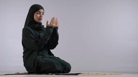 ايماءات تدل على التوسل والتضرع، الدعاء والطلب والتمني من الله، الإجتهاد في العبادة والحفاظ على الصلوات، بورتريه لامراة عربية سعودية خليجية ترتدي الحجاب والعباءة السوداء تجلس على سجادة الصلاة ترفع يديها وتدعو الله، خلفية ملونه