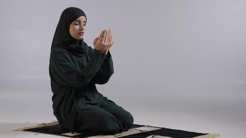 الدعاء والطلب والتمني من الله، ايماءات تدل على التوسل والتضرع، الإجتهاد في العبادة والحفاظ على الصلوات، بورتريه لامراة عربية سعودية خليجية ترتدي الحجاب والعباءة  السوداء تجلس على سجادة الصلاة ترفع يديها وتدعو الله، خلفية ملونه