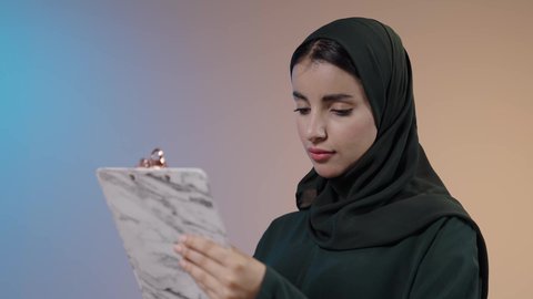 مفهوم التحضير ومراجعة البحوث العلمية ، بورتريه مقرب لشابة عربية خليجيه سعودية محجبه ترتدي العباءة تحمل بيدها حافظة أوراق بيضاء للإستطلاع على المشاريع والأعمال و قراءة الملاحظات، النظر الى شيء ما، ممارسة هواية القراءة ، خلفية ملونة