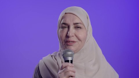 ممارسة موهبة الغناء، تسجيل مقطع صوتي غنائي،  تصوير لامراه عربيه خليجيه سعوديه كبيره بالسن ترتدي العباءه والحجاب تمسك بيدها المايكروفون ،تغني بإيماءات الحماس والسرور، تعابير الاندماج والتفاعل مع الموسيقي، تقديم العروض الصوتية