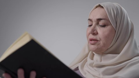 Acquiring knowledge and information, spending free time educating and learning, reading with gestures of contemplation and concentration, a picture of an elderly Saudi Arabian Gulf woman wearing an abaya and hijab sitting on a chair, practicing her hobby of reading useful books in her free time, the concept of literacy.  A picture of an elderly Saudi Arabian Gulf woman wearing an abaya and hijab sitting at a desk.