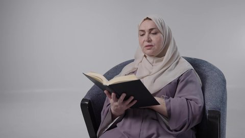 A close-up of an elderly Saudi Arabian Gulf woman wearing an abaya and hijab sitting on a chair. She enjoys reading useful books in her free time. The concept of literacy is to spend one's free time educating and learning, gaining knowledge and information, and reading with gestures of contemplation and concentration.