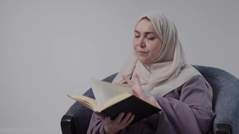 The concept of literacy, spending free time educating and learning, and acquiring knowledge and information. A close-up of an elderly Saudi Arabian Gulf woman wearing an abaya and hijab sitting on a chair, practicing her hobby of reading useful books in her free time. Reading with gestures of contemplation and concentration.