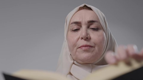 Gaining knowledge and information, spending free time educating and learning, the concept of literacy, a close-up of an elderly Saudi Arabian Gulf woman wearing an abaya and hijab sitting on a chair, practicing her hobby of reading useful books in her free time, reading with gestures of contemplation and concentration.