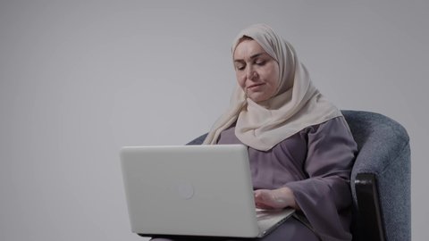 Using modern technologies in administrative work, recruitment in Kuwaiti offices and companies, completing work tasks remotely, integrating advanced technology into daily life, focus, dedication, and hard work. A photograph of an elderly Saudi Arabian Gulf woman wearing an abaya and hijab sitting on a chair and working on a laptop.