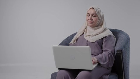 Accomplishing remote work tasks, integrating advanced technology into daily life, using modern technologies in administrative work, hiring in Kuwaiti offices and companies, focus, dedication, and hard work. A photograph of an elderly Saudi Arabian Gulf woman wearing an abaya and hijab sitting on a chair and working on a laptop.