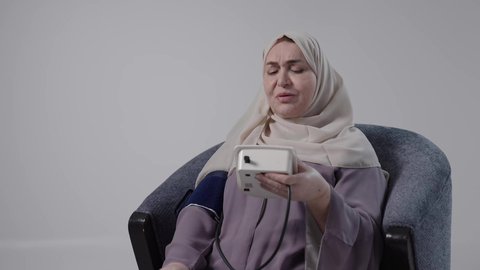 الأجهزة الالكترونية المنزلية الحديثة والمتطورة، الاهتمام والرعاية بالصحة الجسدية, مفهوم الطب والرعاية الصحية, تصوير لامراه عربيه خليجيه سعوديه كبيره بالسن ترتدي العباءة والحجاب تجلس على كرسي ، تحمل جهاز الضغط وتقوم بقياسه ضغط دمها